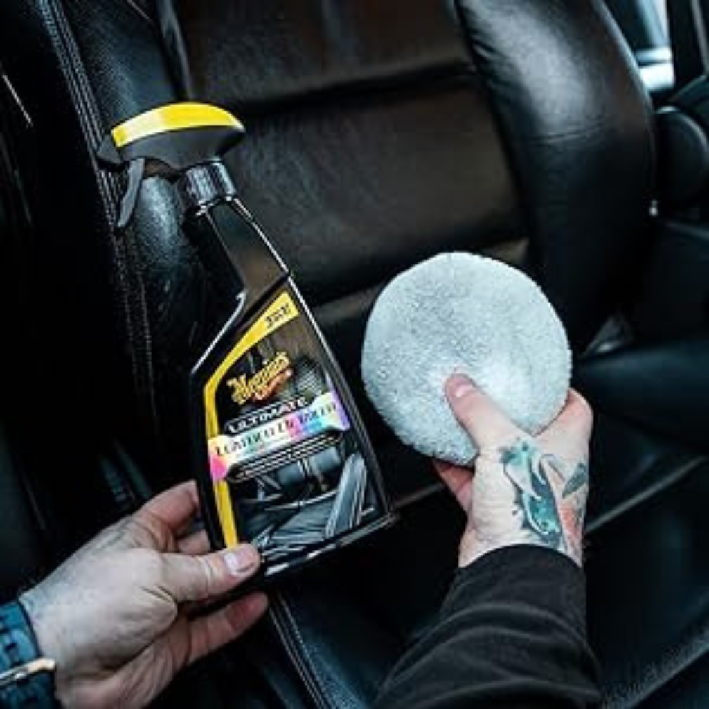 Person holding a bottle of Meguiar's Ultimate Leather Care and a polishing pad inside a car.